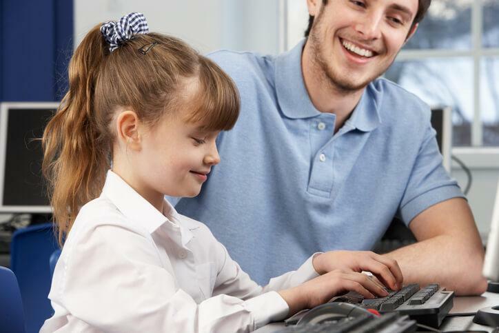 A smiling male educational assistant teaching a pupil how to use the computer after completing his educational assistant training 