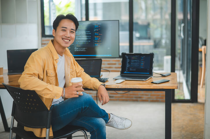 A smiling male programmer working on several computers in an office after completing his web developer training