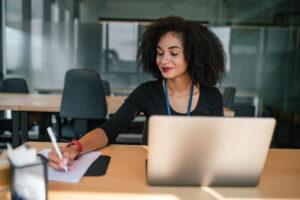 Community service worker writing client case notes at a desk using a laptop and a paper file, demonstrating one of the documentation and case note tips for community service workers